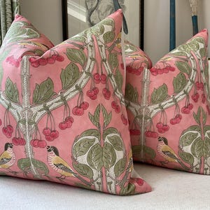 May include: Two pink throw pillows with a floral pattern featuring green leaves, white branches, and red cherries. The pillows have a bird design with yellow and brown accents.