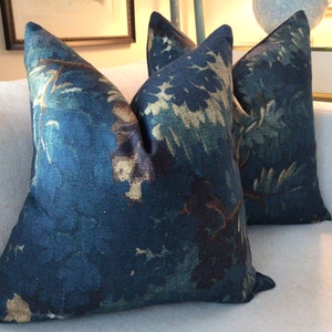 May include: Two decorative throw pillows with a dark blue background and a gold and green floral pattern. The pillows are on a white sofa. The pillows have a textured appearance and are square shaped.