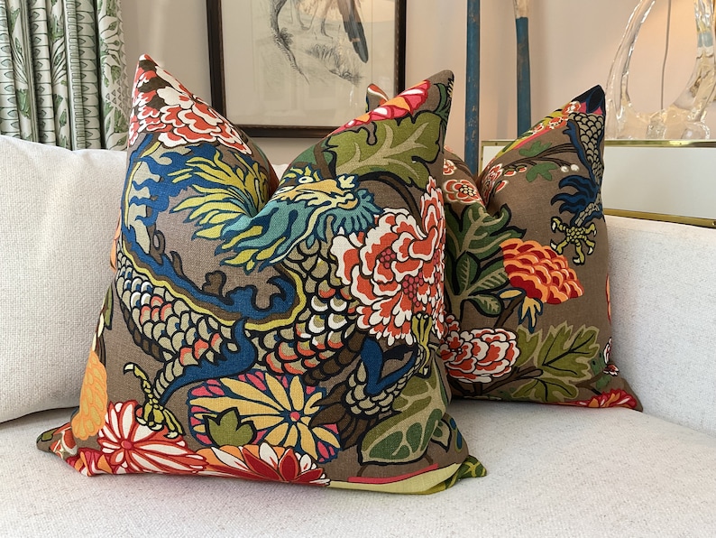 K&ouml;nnte beinhalten: Zwei dekorative Kissen mit braunem Hintergrund und einem bunten Blumen- und Drachenmuster. Die Kissen liegen auf einem wei&szlig;en Sofa.