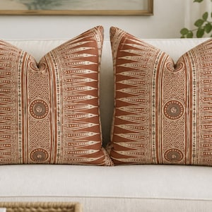 May include: Two square throw pillows featuring a rust-colored fabric with a repeating cream-colored pattern. The design includes vertical bands of pointed triangular shapes and circular motifs with intricate line work centered on each pillow.