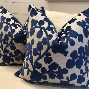 Könnte beinhalten: Zwei dekorative Kissen mit weißem Hintergrund und einem marineblauen Blumenmuster. Die Kissen sind auf einem weißen Sofa platziert. Die Kissen haben ein strukturiertes Aussehen und sind für die Inneneinrichtung konzipiert.