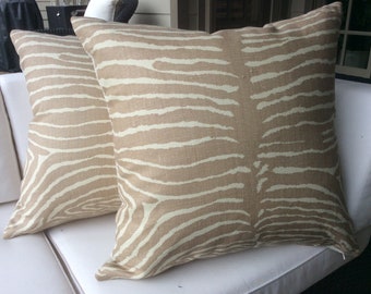 May include: Two beige throw pillows with a white zebra stripe pattern. The pillows are on a white couch.
