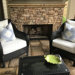May include: Two black wicker chairs with white cushions and blue and white patterned pillows sit in front of a stone fireplace. A glass-top coffee table with a green leaf-shaped bowl sits between the chairs.