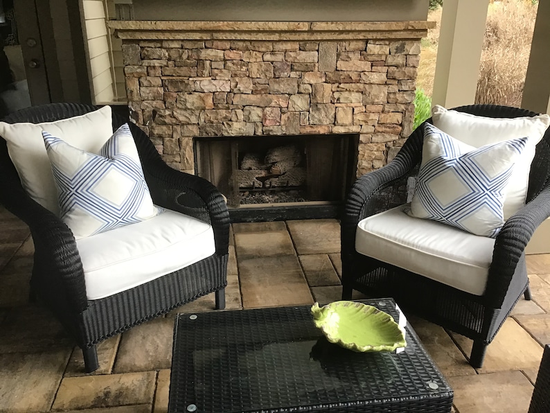 May include: Two black wicker chairs with white cushions and blue and white patterned pillows sit in front of a stone fireplace. A glass-top coffee table with a green leaf-shaped bowl sits between the chairs.