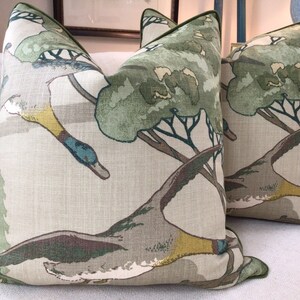 May include: Two green and brown throw pillows with a pattern of flying ducks and trees. The pillows have a light beige background and a green border.