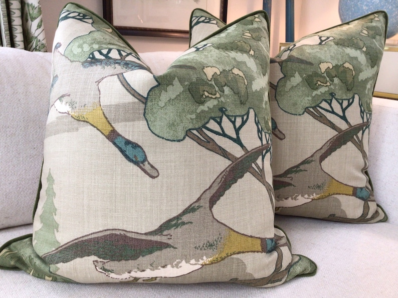 May include: Two green and brown throw pillows with a pattern of flying ducks and trees. The pillows have a light beige background and a green border.