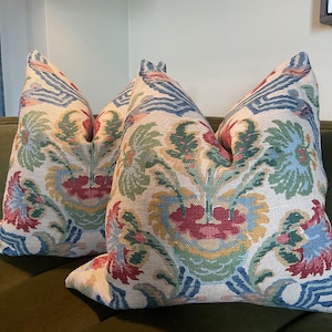 Pode incluir: Duas almofadas decorativas com um padrão floral em vermelho, azul, verde e amarelo sobre um fundo creme. As almofadas estão colocadas num sofá de veludo verde, criando um contraste de texturas e cores.