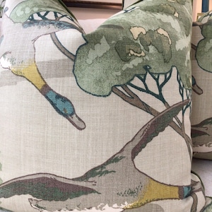 May include: Two green and brown throw pillows with a duck and tree pattern. The pillows are made of a linen fabric.