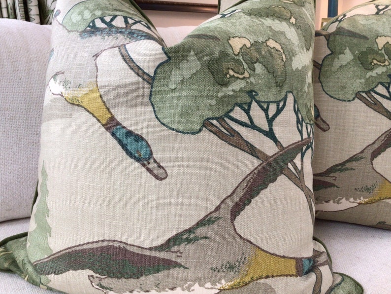 May include: Two green and brown throw pillows with a duck and tree pattern. The pillows are made of a linen fabric.