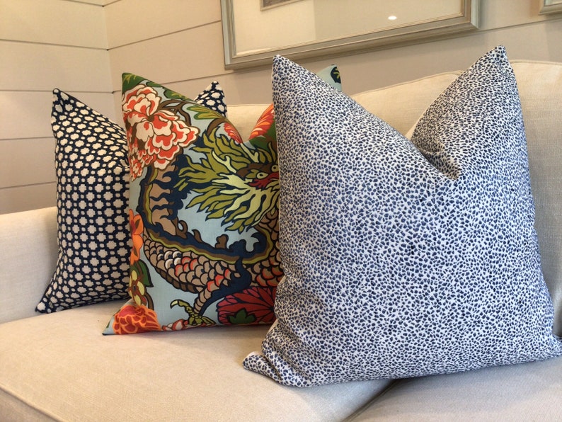 May include: Three decorative throw pillows on a cream colored couch. The pillows are all rectangular and have different patterns. The pillow on the left has a black and white geometric pattern. The middle pillow has a colorful floral pattern with a dragon design. The pillow on the right has a white background with a blue and white speckled pattern.