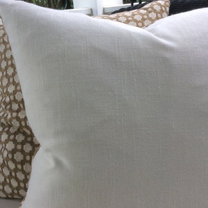 May include: A white linen pillow with a subtle texture. The pillow is placed on top of a brown patterned pillow.
