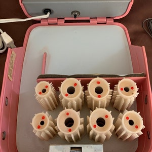 Op de afbeelding: Roze vintage elektrische warmte rollers in een draagtas. De tas is open en de rollers zitten erin. De rollers zijn wit met rode stippen aan de bovenkant.