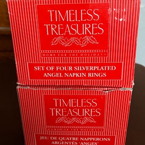Puede incluir: Dos cajas rojas con rayas blancas y letras doradas. La caja superior dice "TIMELESS TREASURES HOME FOR THE HOLIDAYS SET OF FOUR SILVERPLATED ANGEL NAPKIN RINGS". La caja inferior dice "TIMELESS TREASURES HOME FOR THE HOLIDAYS JEU DE QUATRE NAPPERONS ARGENT&Eacute;S 'ANGES'".