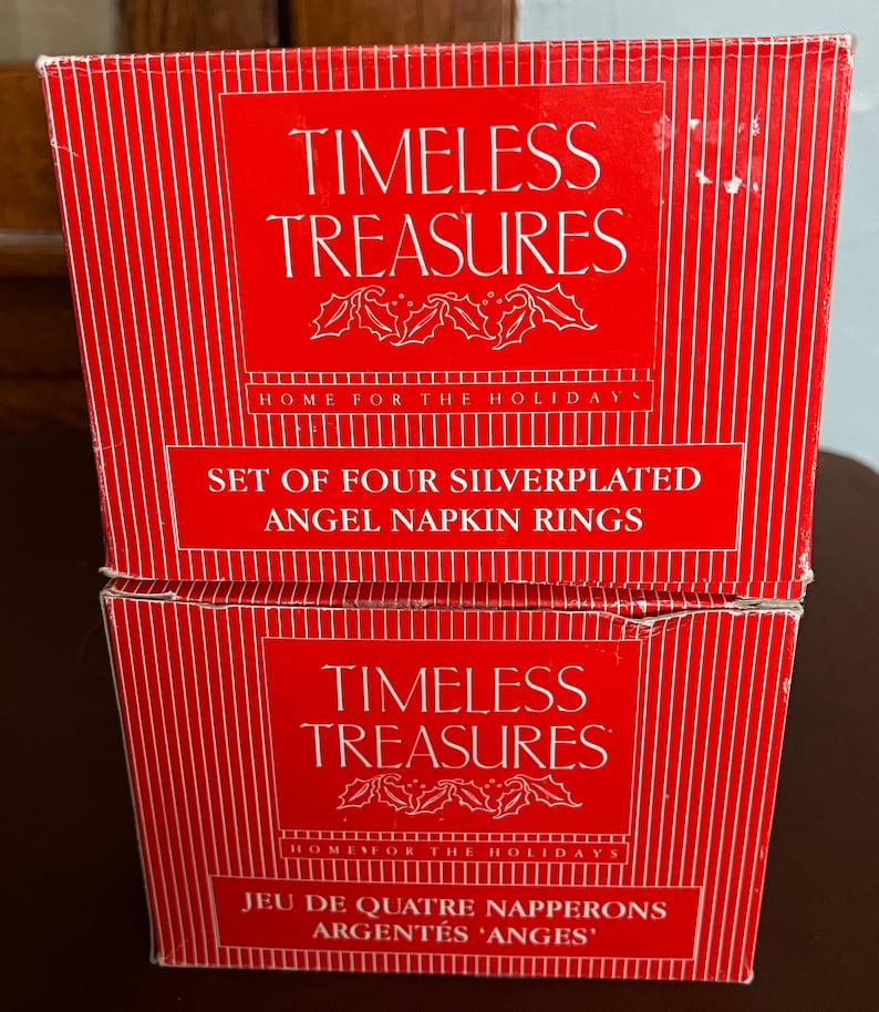Puede incluir: Dos cajas rojas con rayas blancas y letras doradas. La caja superior dice "TIMELESS TREASURES HOME FOR THE HOLIDAYS SET OF FOUR SILVERPLATED ANGEL NAPKIN RINGS". La caja inferior dice "TIMELESS TREASURES HOME FOR THE HOLIDAYS JEU DE QUATRE NAPPERONS ARGENT&Eacute;S 'ANGES'".
