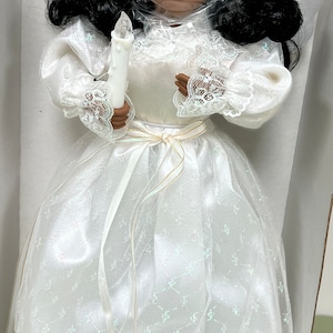 May include: A white porcelain angel doll with black hair and a white dress. The angel is holding a white candle and has a gold ribbon around her waist. The doll is in a white box with a clear plastic cover.