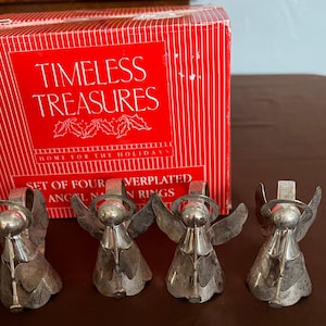 Puede incluir: Conjunto de cuatro anillos de servilleta de &aacute;ngel plateados en una caja roja con las palabras "Timeless Treasures" y "Home for the Holidays". La caja tambi&eacute;n dice "Set of Four Silverplated Angel Napkin Rings".