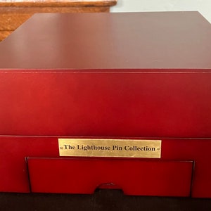Puede incluir: Una caja de madera roja con una placa dorada que dice "The Lighthouse Pin Collection".