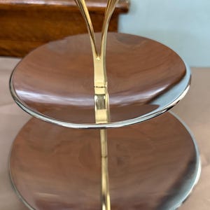 Könnte beinhalten: Zweistöckiges Serviertablett mit goldfarbenem Griff und Rand. Die Tabletts sind silberfarben mit braunem Innenraum. Der Griff hat ein dekoratives Design. Das Tablett ist wahrscheinlich zum Servieren von Desserts oder Vorspeisen gedacht.