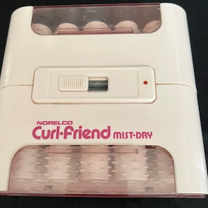 Op de afbeelding: Een witte en roze elektrische krultang met de merknaam "Norelco Curl-Friend MIST-DRY" gedrukt op de voorkant. De krultang heeft een transparante plastic bovenkant met zes roze rollers die zichtbaar zijn in het interieur.
