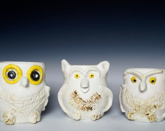 Op de afbeelding: Drie witte keramische uilbeeldjes met gele ogen en zwarte pupillen. De uilen hebben gedetailleerde kenmerken, waaronder veren en snavels.