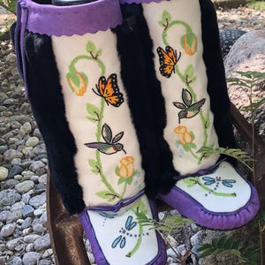 May include: A pair of handcrafted, high-top moccasin boots. The boots feature a black fur shaft with a purple suede cuff. The central panel is cream-colored, adorned with embroidered butterflies, hummingbirds, and floral designs. The soles are white with dragonfly embroidery and purple trim.