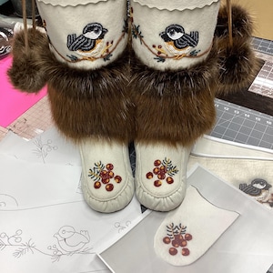 Puede incluir: Un par de mocasines de cuero blanco con ribete de piel marrón y diseños de cuentas de pájaros y bayas. Los mocasines están sobre una superficie blanca con un patrón de pájaros y bayas.