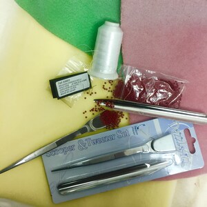 May include: A close-up of beading supplies including red seed beads, a spool of white thread, a pair of tweezers, a bead scooper, and a package of beading needles. The text on the package of needles reads "743 23031 Beading Needle No.11 Gold Eye Size 0.6MM x 52.5 MM Japanese Quality Carp Made in Japan".