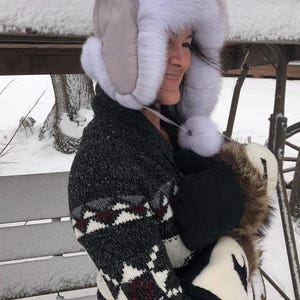 May include: A woman wearing a white fur hat with a gray brim and a black and white patterned sweater. She is holding a white fur muff with a brown fur trim.