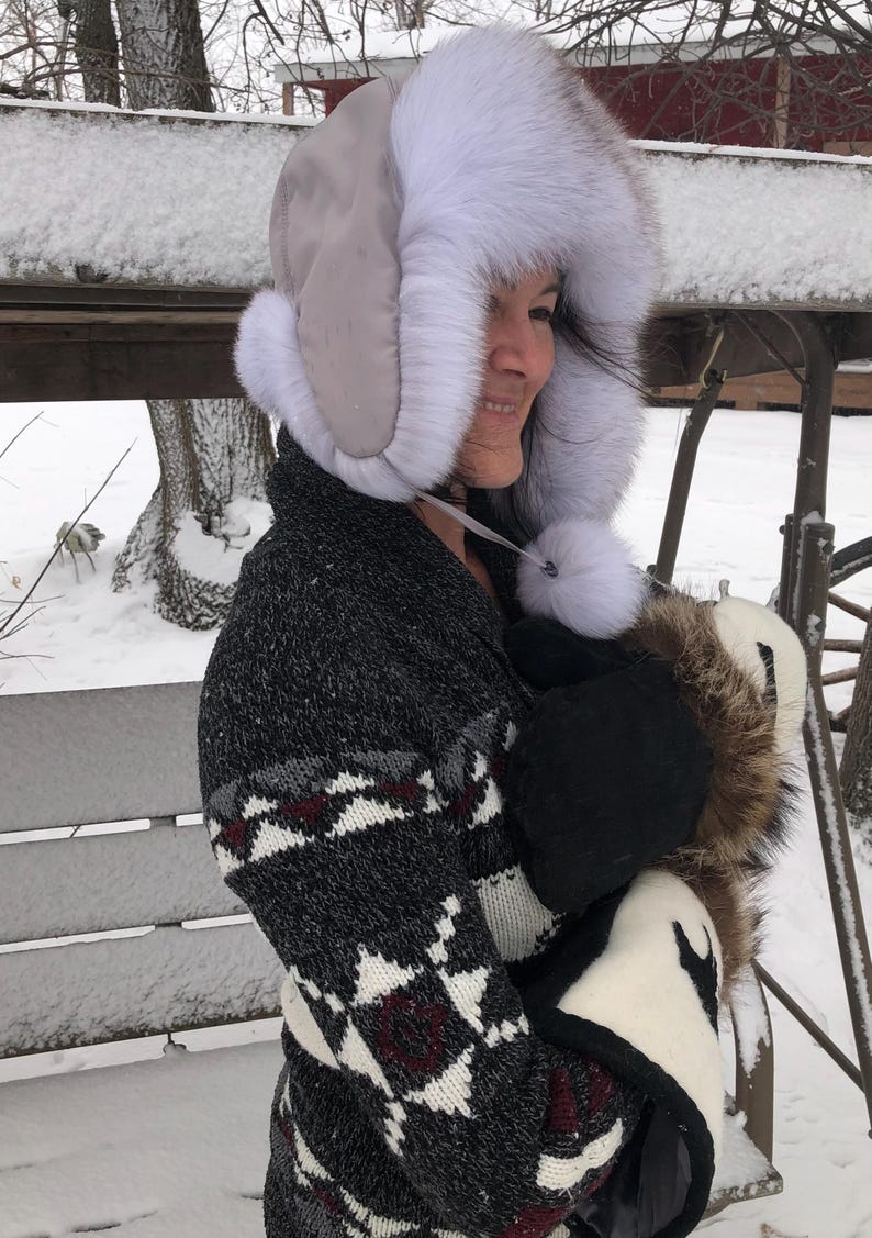 May include: A woman wearing a white fur hat with a gray brim and a black and white patterned sweater. She is holding a white fur muff with a brown fur trim.