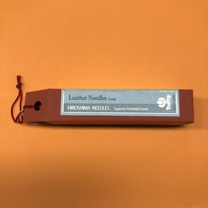 May include: A brown box with a white label that reads "Leather Needles Long" and "HIROSHIMA NEEDLES Superior Polished Finish". The box has a red string attached to it.
