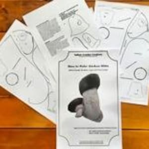 May include: Printed paper patterns and instructions for creating a mushroom-shaped item. The main image on the instructions shows a detailed illustration of the finished product, featuring a beige stem and a dark brown cap. The papers are arranged on a wooden surface.