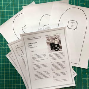 Può includere: Un set di modelli di carta bianca per realizzare guanti senza dita. I modelli sono etichettati con le dimensioni e le istruzioni di taglio. I modelli sono su un tappetino da taglio verde.