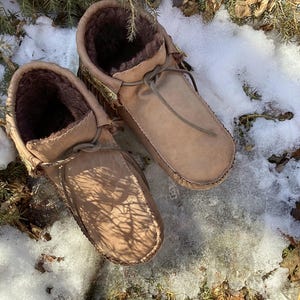 Peut inclure: Une paire de mocassins en cuir marron avec des détails en franges et une doublure en fourrure. Les mocassins sont posés sur un sol enneigé avec de la végétation verte.