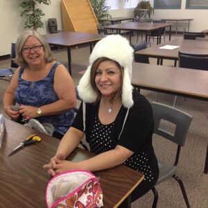 May include: Two women sitting at a table in a classroom. One woman is wearing a white fur hat and a black and white polka dot shirt. The other woman is wearing a blue tank top and has short brown hair. There is a pink and white patterned makeup bag on the table.