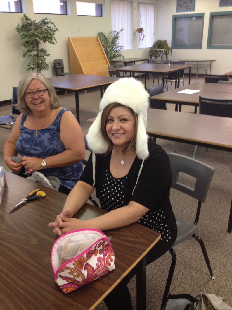 May include: Two women sitting at a table in a classroom. One woman is wearing a white fur hat and a black and white polka dot shirt. The other woman is wearing a blue tank top and has short brown hair. There is a pink and white patterned makeup bag on the table.