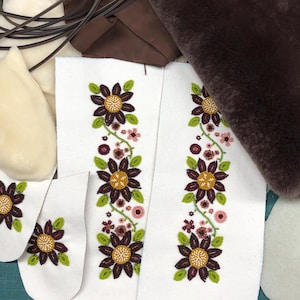 May include: Close-up of white fabric pieces with embroidered floral designs in brown, green, and yellow. Additional components include brown leather, cream-coloured fleece, and a dark brown fur-like material, suggesting a craft project.
