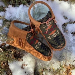 Peut inclure: Une paire de mocassins en daim marron avec une doublure à motif géométrique vert, noir et orange. Les mocassins ont des franges et sont posés sur une surface enneigée.
