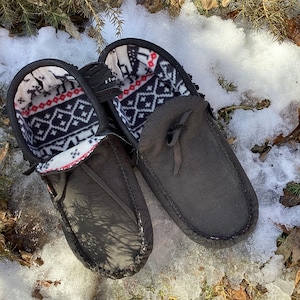 Peut inclure: Une paire de mocassins en cuir noir avec une doublure en polaire à motifs géométriques. Les mocassins sont posés sur un lit de neige et d'aiguilles de pin.