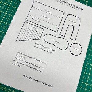 May include: A paper pattern for ornamental mini mukluks, featuring various cut shapes labeled 'Ribbon', '1 inch FUR', 'Muk Upper', 'FRINGE', 'Top FOOT', 'FOOT', and 'Top cut'. The pattern includes text from Ambers Leather Creations.