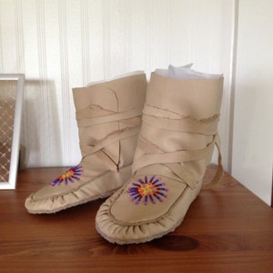 May include: A pair of beige leather moccasins with beaded embellishments. The moccasins have a decorative fringe around the top and are tied with laces.