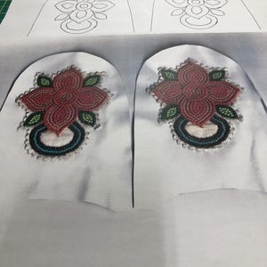 Könnte beinhalten: Zwei weiße Stoffstücke mit einem floralen Design. Das Design ist auf einem Stück in Schwarz umrandet und auf dem anderen mit roten, grünen und blauen Perlen besetzt.