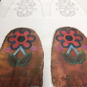 Könnte beinhalten: Zwei braune Ledermokassins mit floraler Perlenarbeit. Jeder Mokassin hat eine rote Blume mit schwarzem Zentrum, blauem Stiel und grünen Blättern. Im Hintergrund befindet sich ein Papier mit einer Blumenkontur.