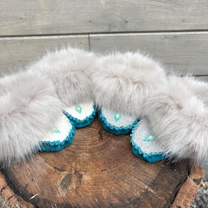 May include: Four pairs of grey fur slippers with teal trim and white and grey embroidered details. The slippers are displayed on a wooden surface, showcasing their soft texture and design.