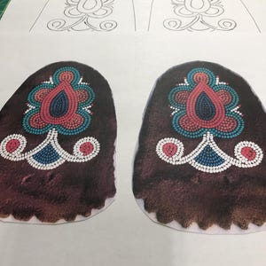 Könnte beinhalten: Zwei braune Ledermokassins mit weißen, blauen und rosa Perlenmustern. Die Mokassins liegen auf einem weißen Blatt Papier mit einer Bleistiftumrandung des Perlenmusters.