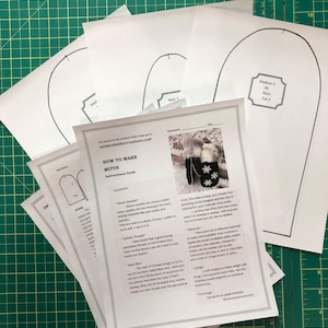 Puede incluir: Un conjunto de patrones de papel blanco para hacer mitones. Los patrones están etiquetados con tamaños e instrucciones de corte. Los patrones están sobre una estera de corte verde.