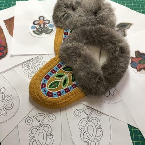 Könnte beinhalten: Nahaufnahme eines Paares Mokassins mit Pelzbesatz. Die Mokassins sind aus Leder und haben aufwendige Perlenmuster. Auf dem Tisch liegen mehrere Papiermuster mit ähnlichen Designs.