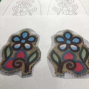 Könnte beinhalten: Zwei Nahaufnahmen von Perlen-Blumenmustern. Die Muster sind auf einem braunen Stoffgrund und haben blaue, rote, weiße und braune Perlen. Die Muster sind von einem weißen Hintergrund umgeben.