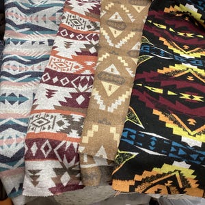 May include: Four rolls of patterned fabric in various colours. The fabrics feature geometric designs in shades of blue, pink, brown, and black. The patterns include diamonds, triangles, and zigzags, creating a Southwestern or tribal aesthetic. The rolls are stacked together.