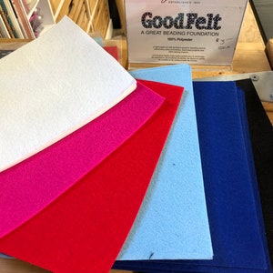 Peut inclure: Une pile de feuilles de feutre de différentes couleurs, dont blanc, rose, rouge, bleu clair, bleu foncé et noir. Le feutre est étiqueté "GoodFelt, A Great Beading Foundation, 100% Polyester".