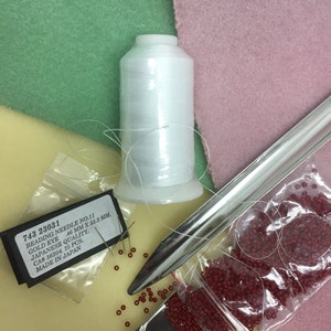 May include: A close-up of beading supplies including a spool of white thread, a metal beading tool, a small bag of red beads, and a package of beading needles labelled "743 23031 Beading Needle No. 11 Gold Eye 46 mm x 33.3 mm. Japanese Quality. CA# 36204. 25 Pcs. Made in Japan."